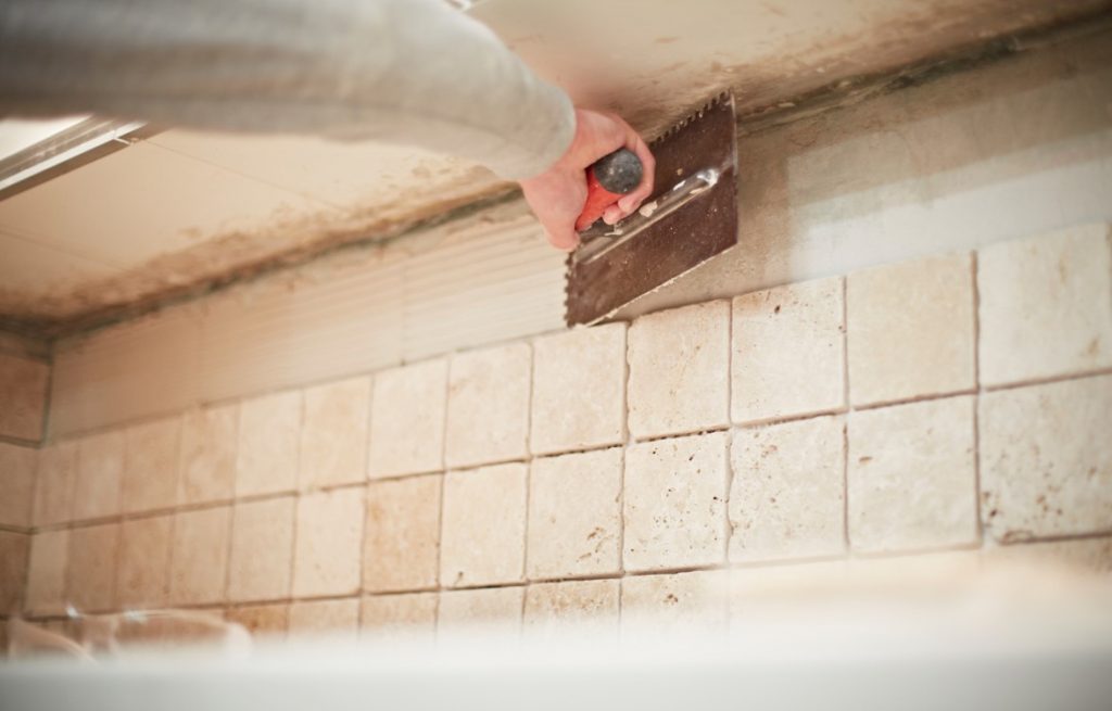 Hand holding a tiling tool, applying adhesive to a wall that they are fixing tiles to. Two rows of tiles are already fixed.