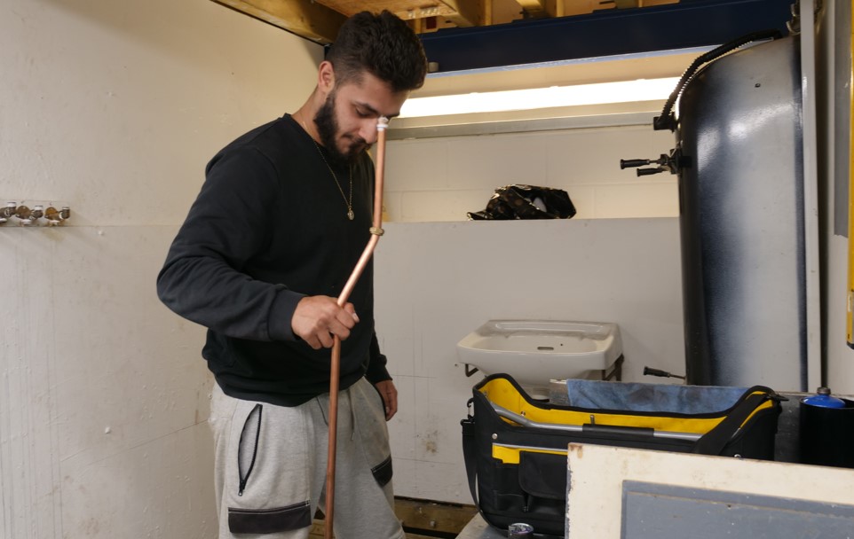Student in Able Skills plumbing centre completing their plumbing training. Student is holding a copper pipe, standing next to an open tool bag in a bathroom area.