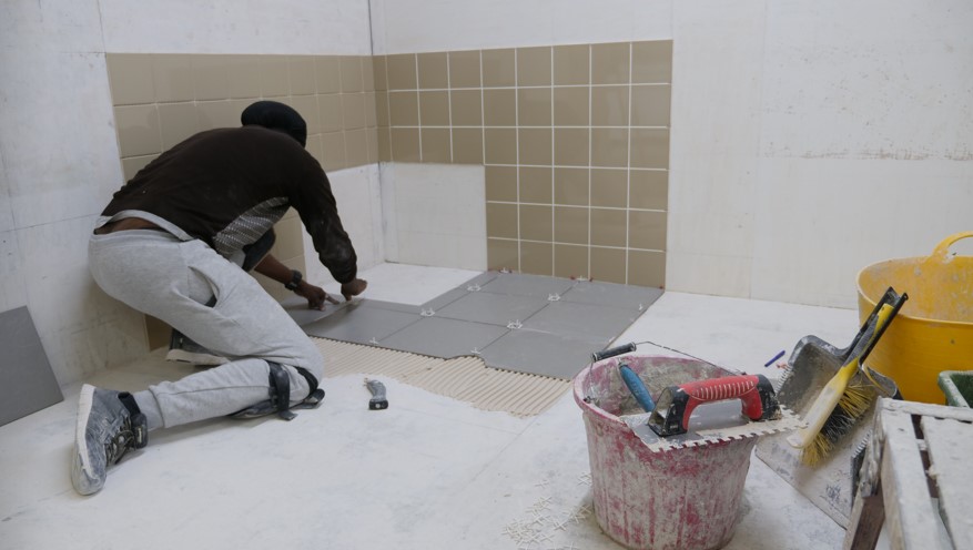 Student at Able Skills Construction training is working on tiling a bathroom area using grey floor tile and brown wall tiles. They are applying adhesive and using tile spacers, and wearing knee pads as they are working on the floor. 