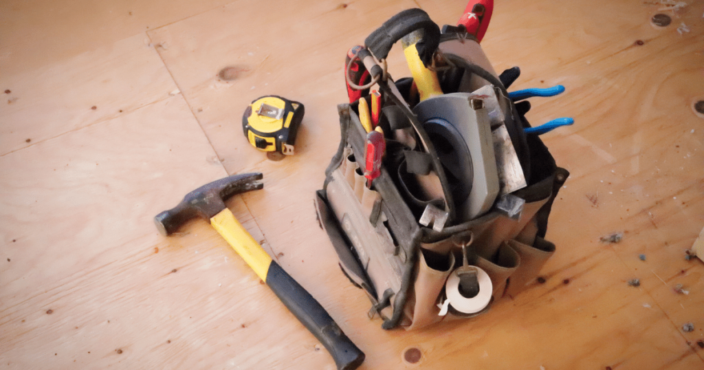 A hammer is on the wooden floor, next to a tape measure and a tool bag with a selection of tools typical for someone working in the construction industry