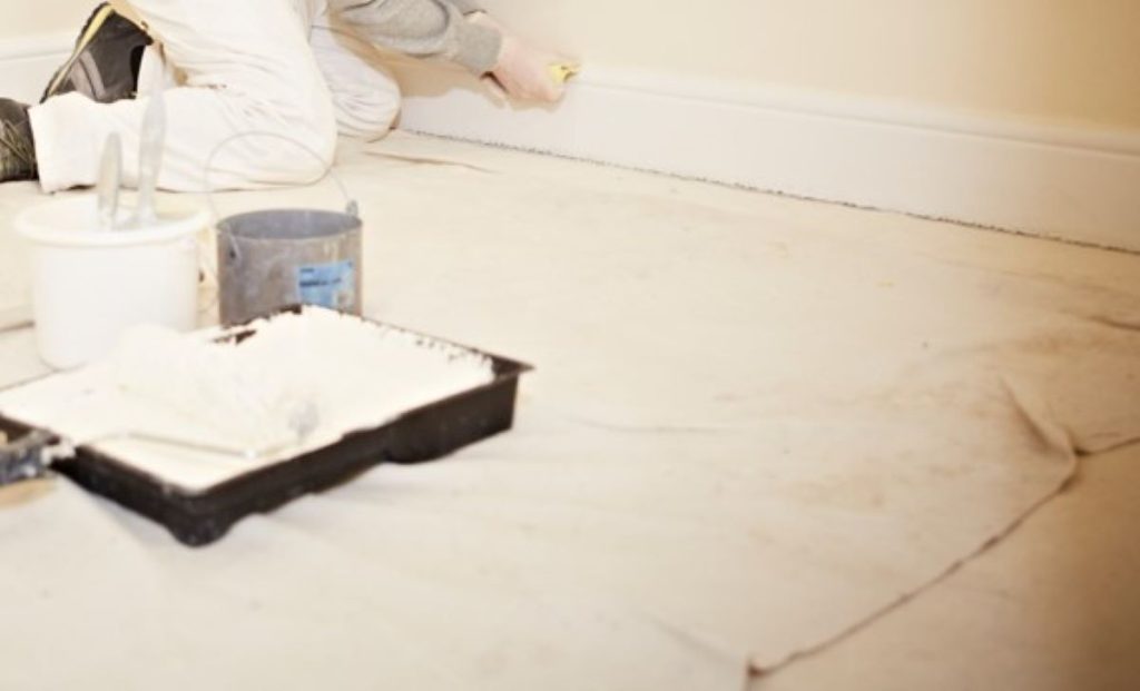 Dust sheet on a floor with a paint roller tray and a person kneeling is sanding a skirting board