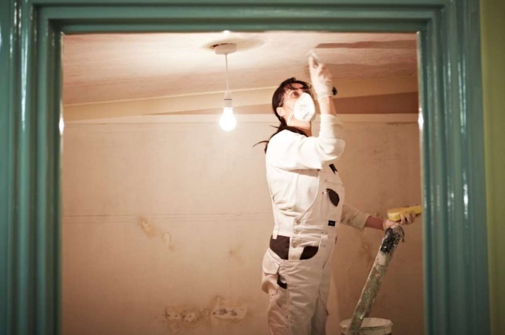 Woman on a step-ladder mis-coating a freshly plastered ceiling. She is wearing white painting overalls and a dust mask