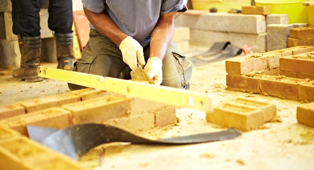 Person kneeling down applying mortar to bricks. In front of them is a 2-course brick wall with a level on top. 
