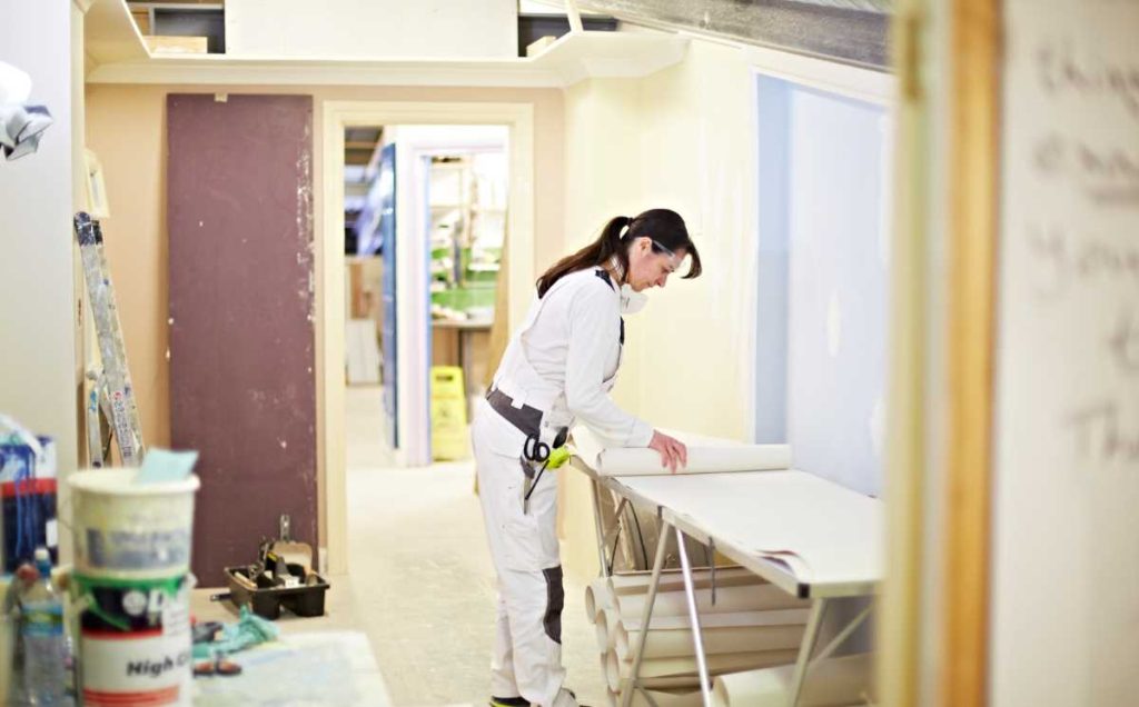 Woman leaning over a wallpaper table unrolling wallpaper. She is surrounded by paint pots and tools. 