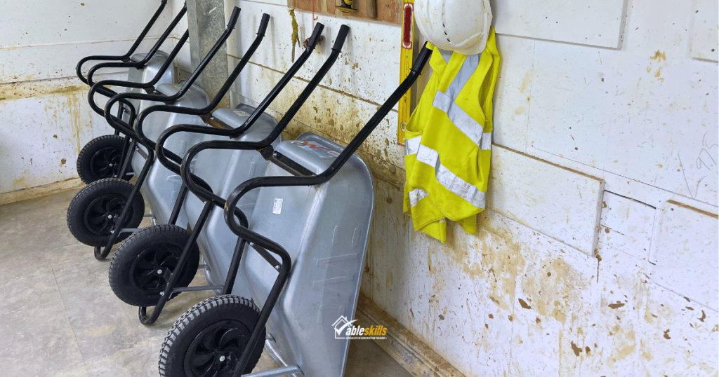 Wheelbarrows leaning against a wall, a hi-viz jacket and hard hat are hanging next to them