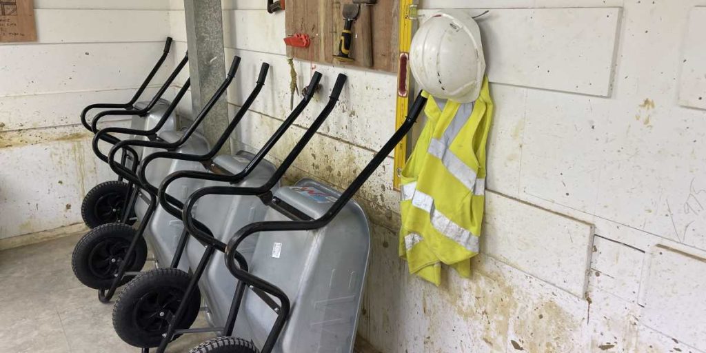 Wheel barrows leaning against a wall next to a hi-viz jacket and hard hat