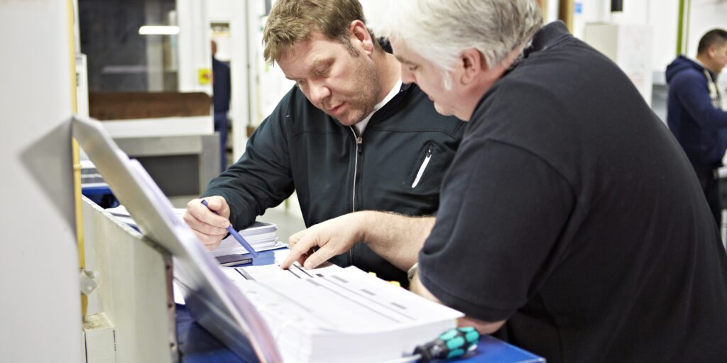 Two men are reviewing documents together in a workshop or training environment. One man, with short light-colored hair, points at the papers while the other man, with short brown hair, looks closely and holds a pen, appearing to take notes or discuss details. Tools and equipment can be seen in the background, suggesting a technical or vocational setting, possibly related to gas training or certification.