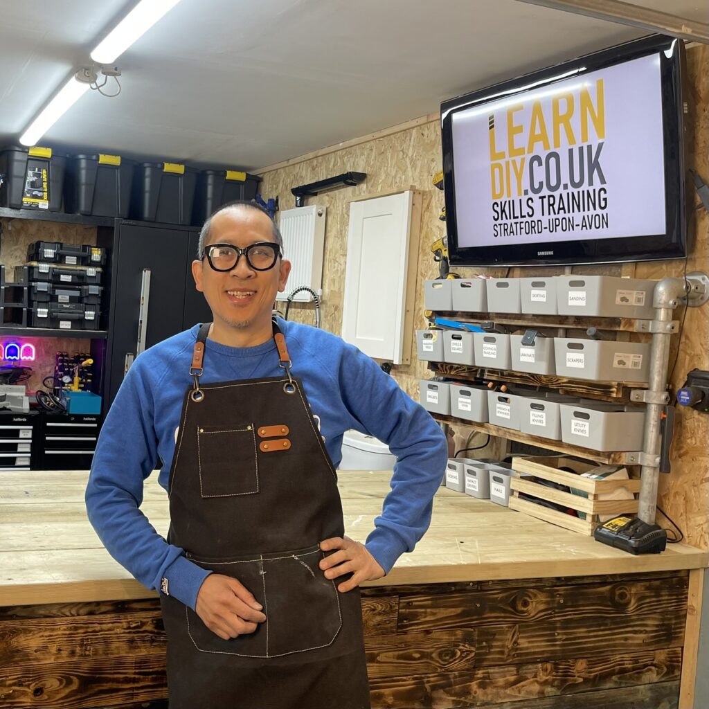 Mike Lok is wearing an apron and a blue sweatshirt. He is looking at the camera smiling, wearing dark framed glasses. Behind him, there is a workbench and various tools, storage boxes and equipment neatly arranged. A monitor on the wall has the words Learn DIY Skills Training on display.