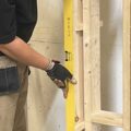 A person, wearing work trousers and a tool belt, is holding a yellow level against a wooden structure, possibly the framework for a wall.