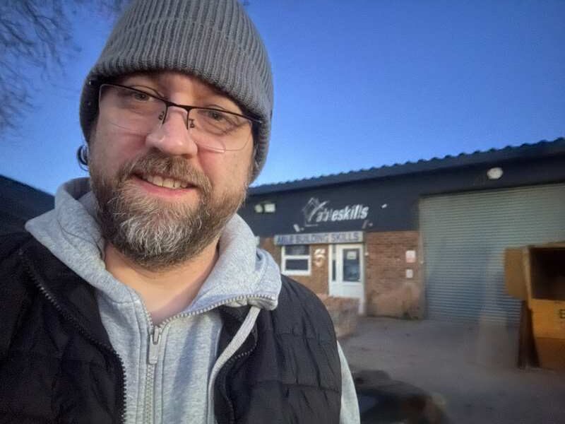 Paul is wearing a grey beanie and glasses. He's standing in front of the Able Skills construction training centre