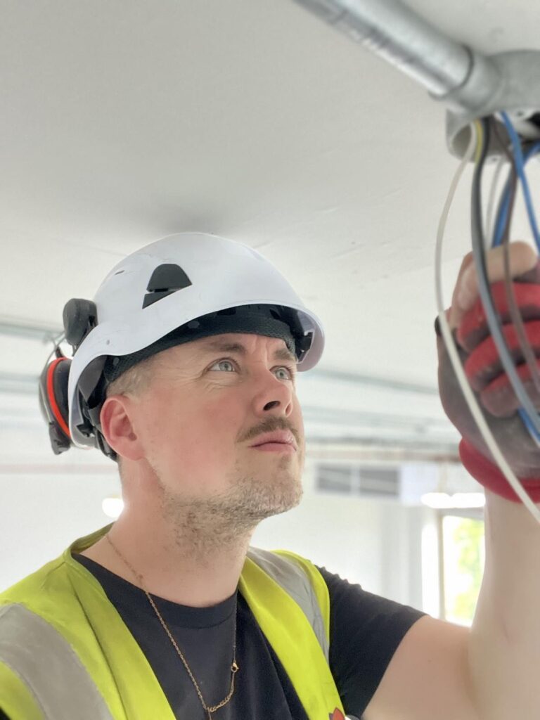 Scott wearing a safety hat and hi-vis vest. he is running wiring through mental trunking.