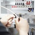 Hands holding wires working on electrical installation. A fuseboard in the back ground