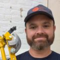 Peter Chapman plumber is facing the camera wearing a cap with a logo and holding a set of pipe benders