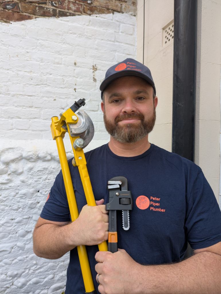 Peter Chapman faces the camera wearing a cap and t-shirt with a plumbing company logo and he's holding a pipe bender and wrench