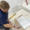 An Able Skills instructor kneels on the floor, demonstrating how to lay floor tiles.